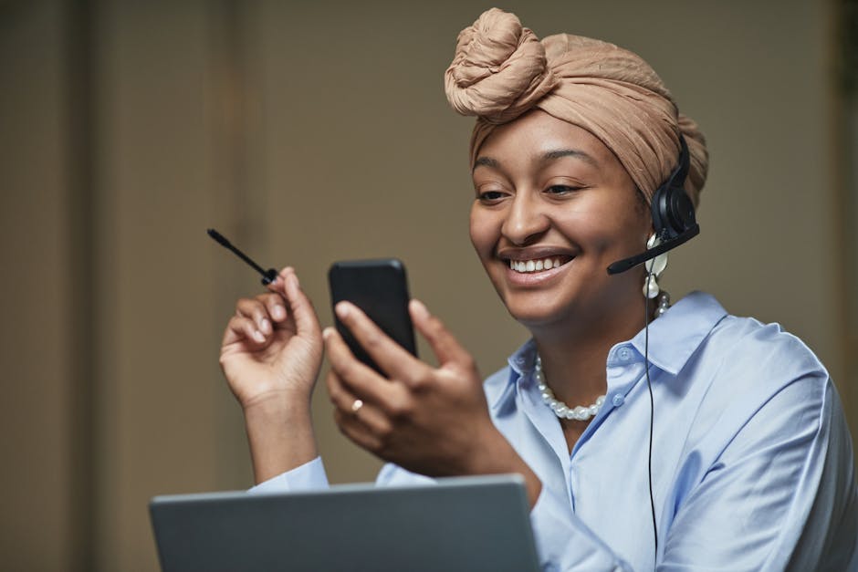 A cheerful woman in a headwrap and headset multitasking between smartphone and laptop in an indoor office setting.