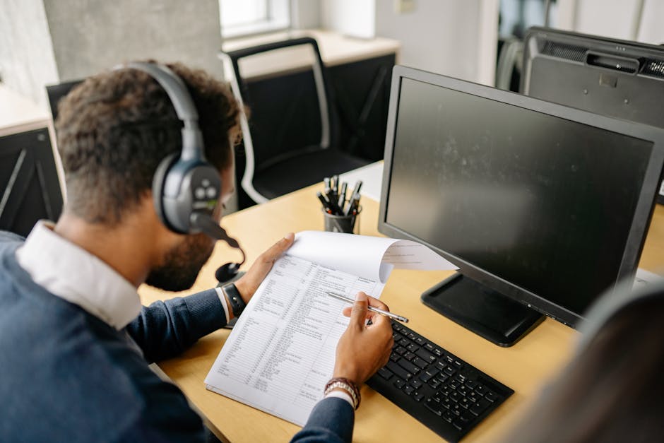 Adult male using a computer and headset, reviewing documents in a modern office setting.